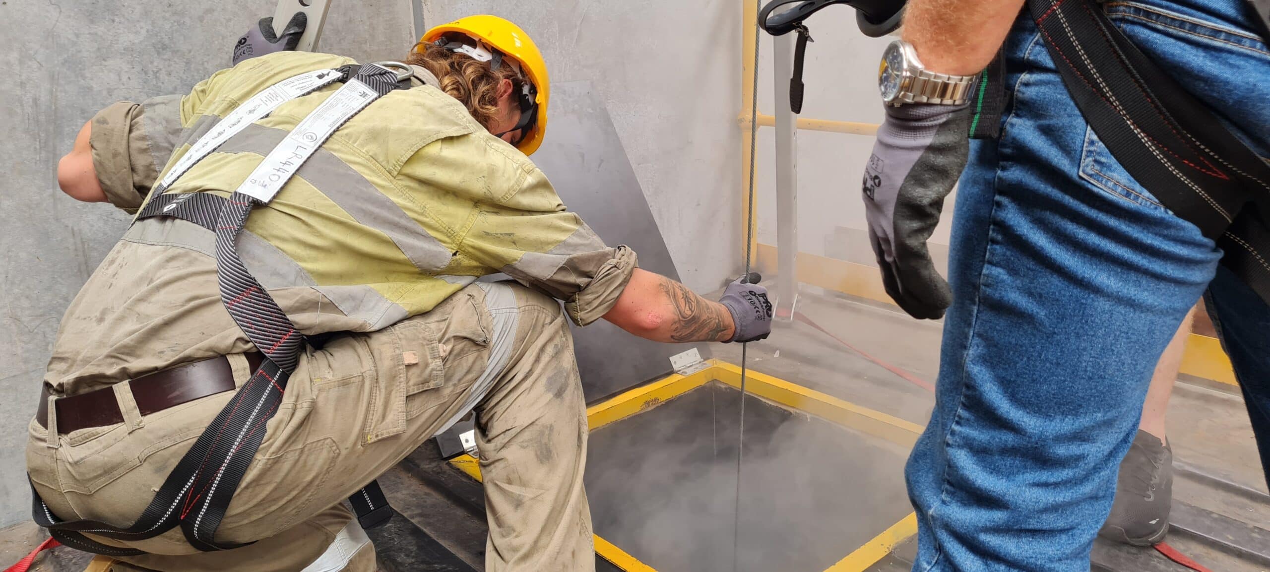 Construction worker in hard hat and harness leans over an open shaft.