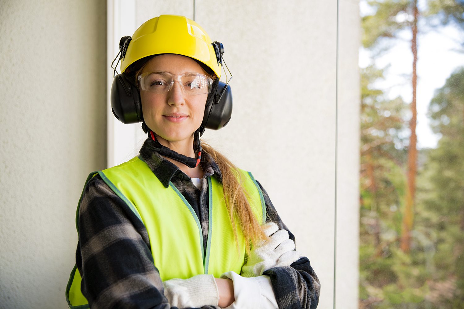 A woman wearing a yellow hard hat, safety glasses, a yellow safety vest over a plaid shirt, and black earmuff-style hearing protection is standing with her arms crossed.